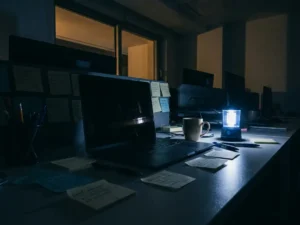 Donker kantoorbureau met uitgevallen stroom, zwart laptopscherm en noodlantaarn die blauw licht werpt op notities en koffiemok.