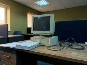 Verouderde desktop-pc met bolle CRT-monitor naast open EU-regelgevingsbinder op modern bureau, tangled kabels, koel tl-licht.